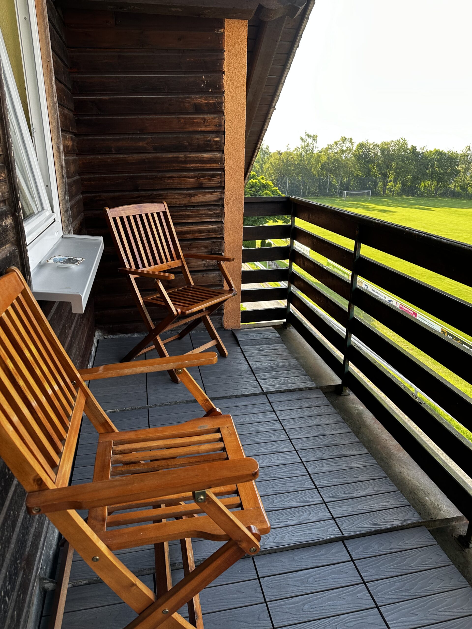 Zwei Holzklappstühle auf einem Balkon mit einem Blick auf eine grüne Fläche.